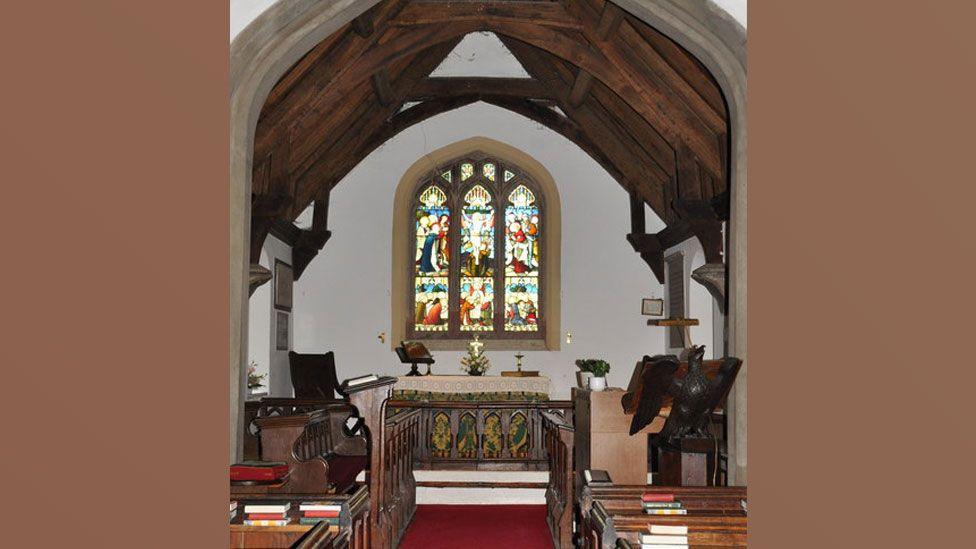 Interior of St Andrew's Church, Greensted, looking towards the altar. It shows an arched stained glass window above a cloth-covered altar. It has white walls and the a pitched timber roof. There is a red carpet on the nave and wooden pews can be seen on either side.
