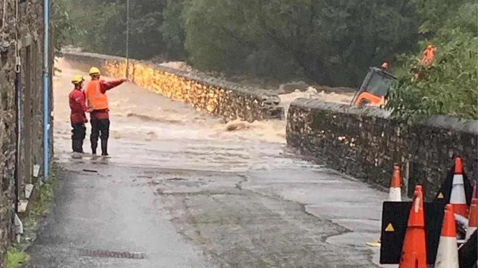 Water gushing out of the River Laxey onto the road through a hole in a stone wall. Men in red and orange hi-vis coats and vests point and an orange digger on its side in the river.