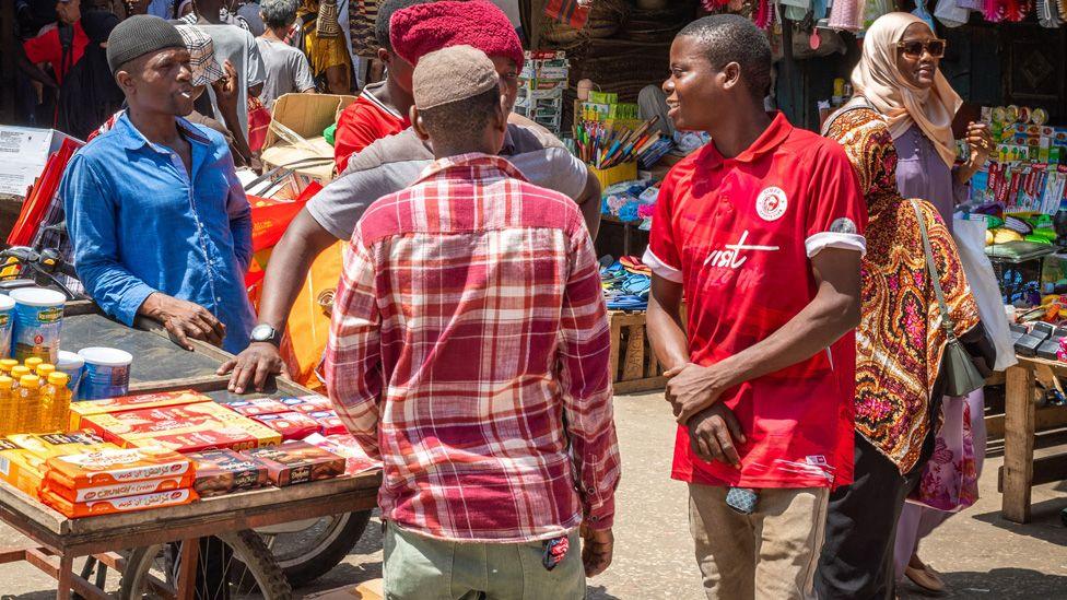 Traders chat at a market in Zanzibar as other shoppers, including a woman in a headscarf and sunglasses, walk past.