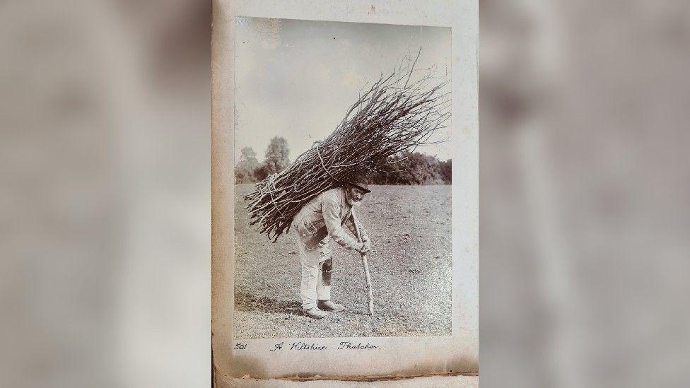 Black and white/sepia image of a man with a beard carrying sticks on his back