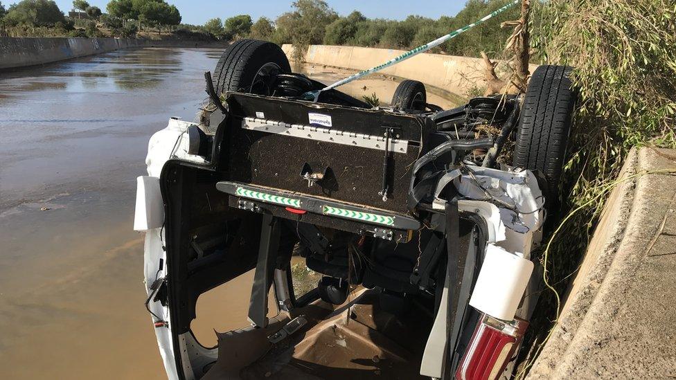 A white SUV-style car is pictured standing on its roof, flipped, in shallow water - it is surrounded by a police cordon