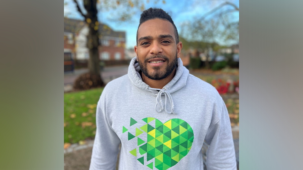 Liam Edwards is wearing a grey hoodie with a green heart mosaic type logo in the centre. In the background is a street with a grass verge