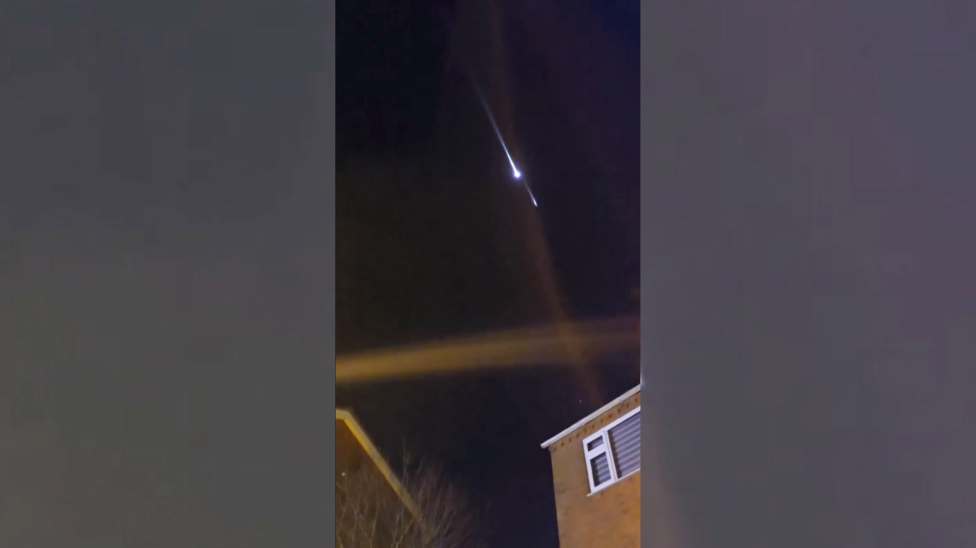 A photos of a bright ball with a bright streak trailing behind travelling through the sky. Corners of houses can be seen at the bottom of the photo.