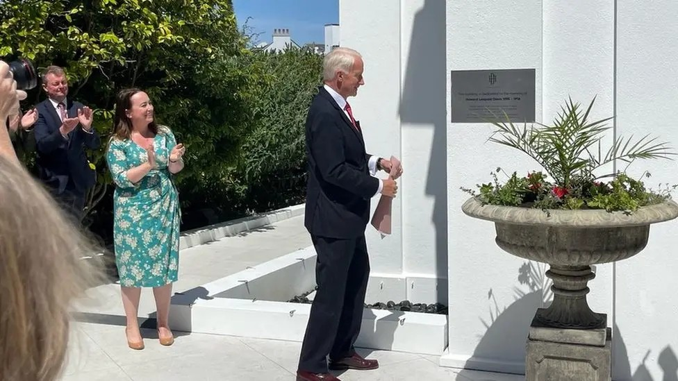 A man with grey hair and a dark suit opens a white building surrounded by plants.