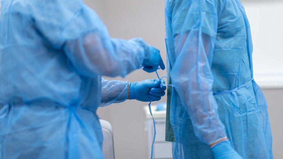 A surgeon in an operating theatre, dressed in full scrubs, helps a colleague, dressed in the same manner, tie his scrubs