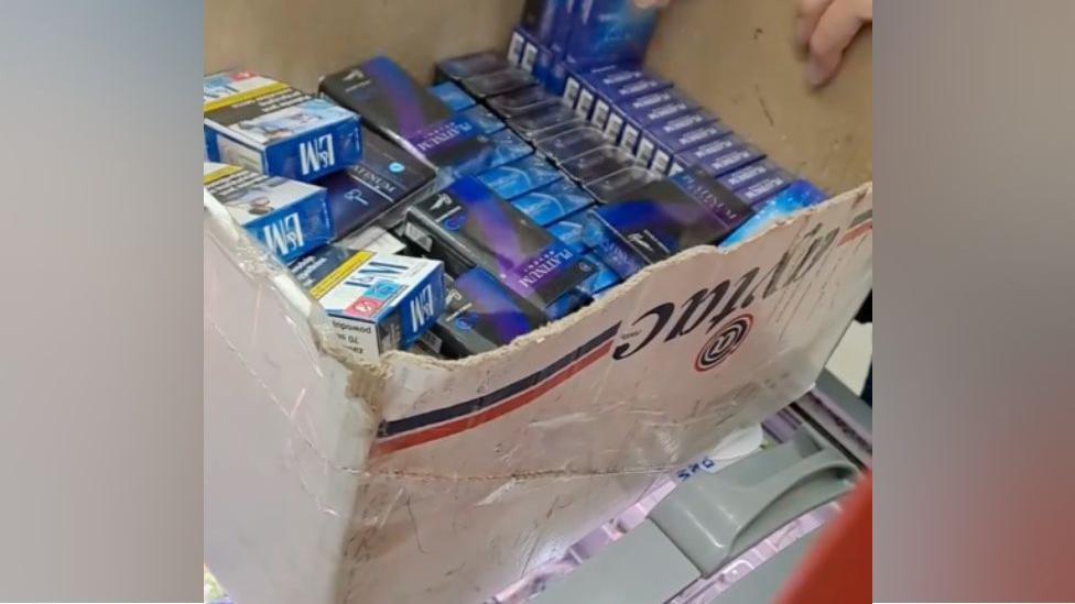 Boxes of medication are in blue packets and sitting in a cardboard box. The boxes have cellophane wrapping on them and are coloured blue, purple, white and yellow.  Two fingers from a hand can be seen on an edge of the box, holding it.