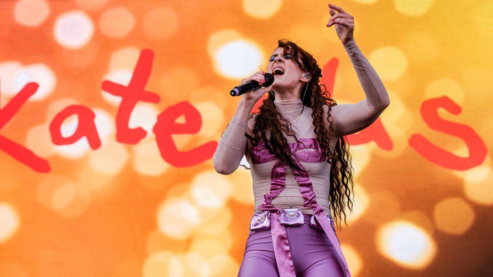 Kate Nash singing on stage in a purple outfit against an orange backdrop with her name on it in giant letters