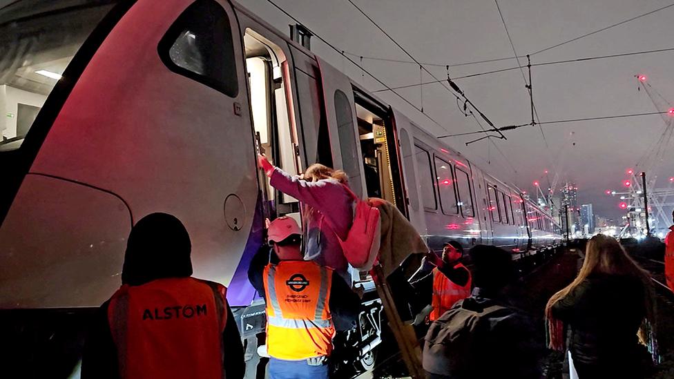 Officials assist passengers to get down from the train, with the train tracks in the background.