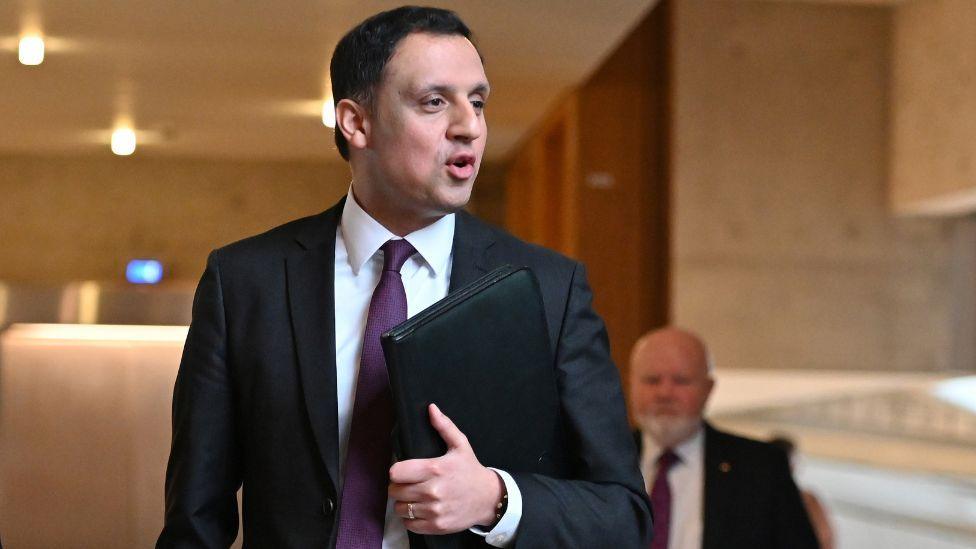 Anas Sarwar, a man with dark hair, photographed in the Scottish Parliament. Visible from about the waist up, he is holding a black A4-sized folder in his left hand and looking to his left. He is wearing a dark suit, white shirt and purple tie. 