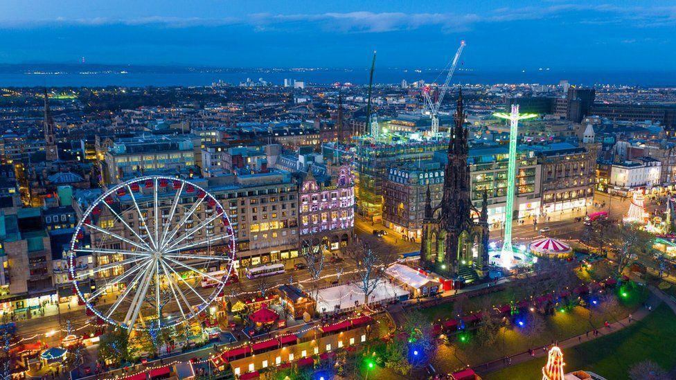 A drone view of Edinburgh city centre. There re lots of lights. The big wheel is on the left of the picture and the Scott Monument is on the right. It is night time. 