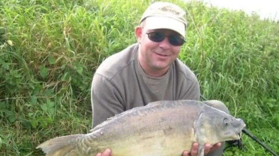 Mr Ricketts again pictured in a khaki outfit holding a large fish. This time he wears dark oval-shaped glasses and a lighter coloured cap. 