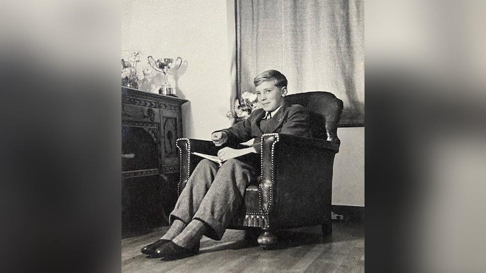 Michael May as a young boy sitting in an armchair next to a trophy