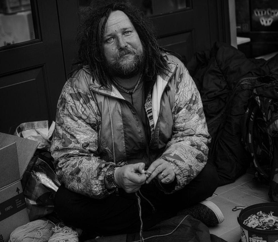 Bearded homeless man sits in doorway making bracelets from string