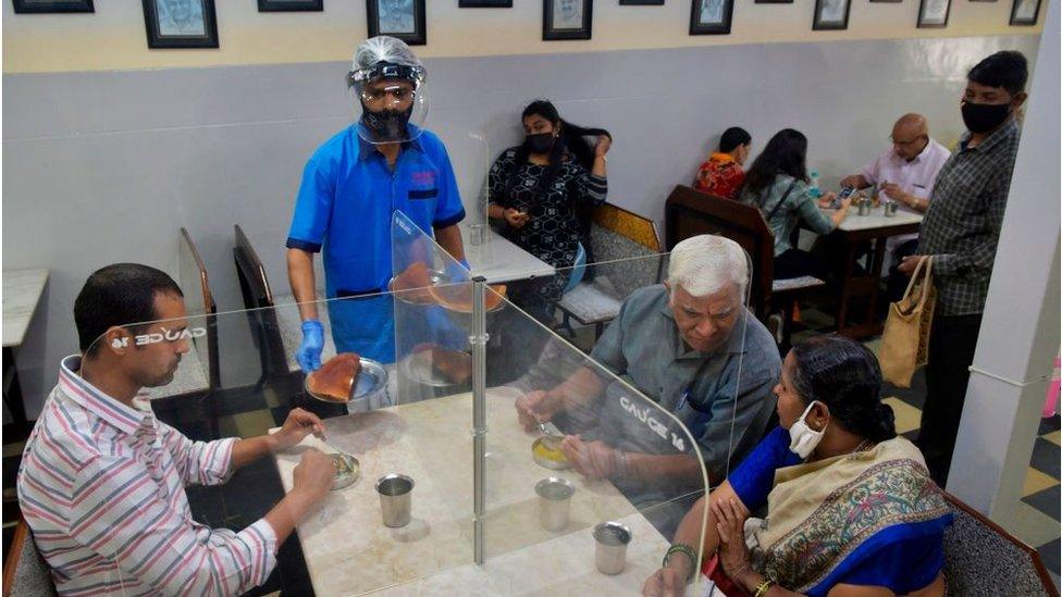 A waiter wearing a face shield and mask serves food to customers sitting in a table divided with transparent panels.