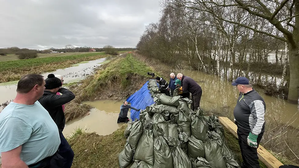 A picture shows a gathering of people on the banks of the Fosse Dyke. The people are piling sand bags into a hole in the bank. Some people are in the water in waders.