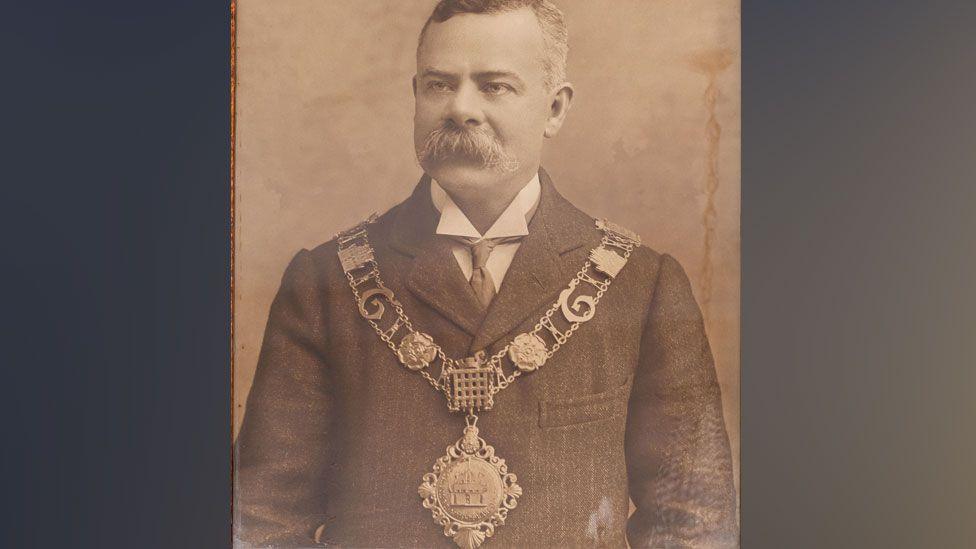 Allan Minns in about 1904, in a sepia-toned black and white photo. He is has short greying hair and a large bristling moustache, covering his upper lip. He is wearing a jacket and underneath a wing-tipped white shirt. Over his jacket is a mayoral chain. It has two rows of links and a large medallion dangling across his stomach