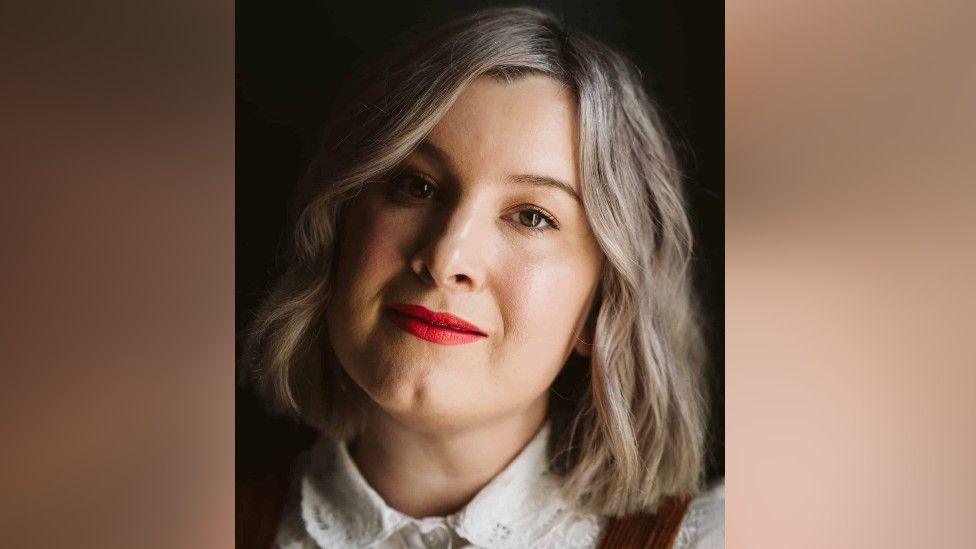 Ellie Gillard is looking into the camera. She is smiling slightly, and is wearing red lipstick. Her hair is blonde and shoulder length.