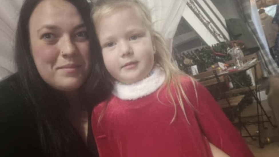 A woman with long dark hair and a nose ring wearing a black top. She has her arm round a young blonde-haired girl who is wearing a red velvet dress with a fluffy white collar. There are tables and chairs behind them.