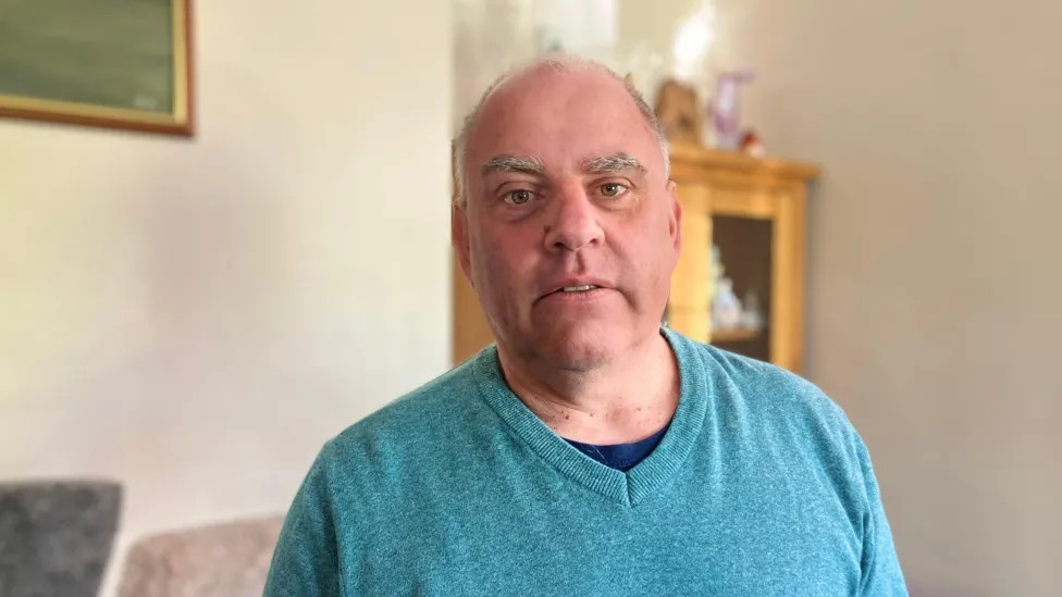 A man stands in his living room. He has grey hair and is bald on top, and wears a blue top.