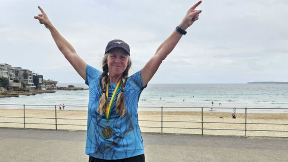 Nikki Love's arms are raised in a victory pose at Bondi Beach. She has long, braided, fair hair and wears a baseball cap, a blue sports top and a medal around her neck.