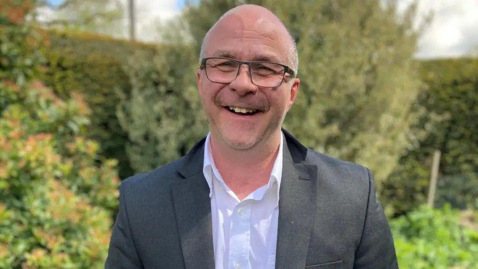 A man with very short white hair and black glasses is smiling at the camera. He has a white shirt on with a black suit blazer. Behind him are green bushes and trees.