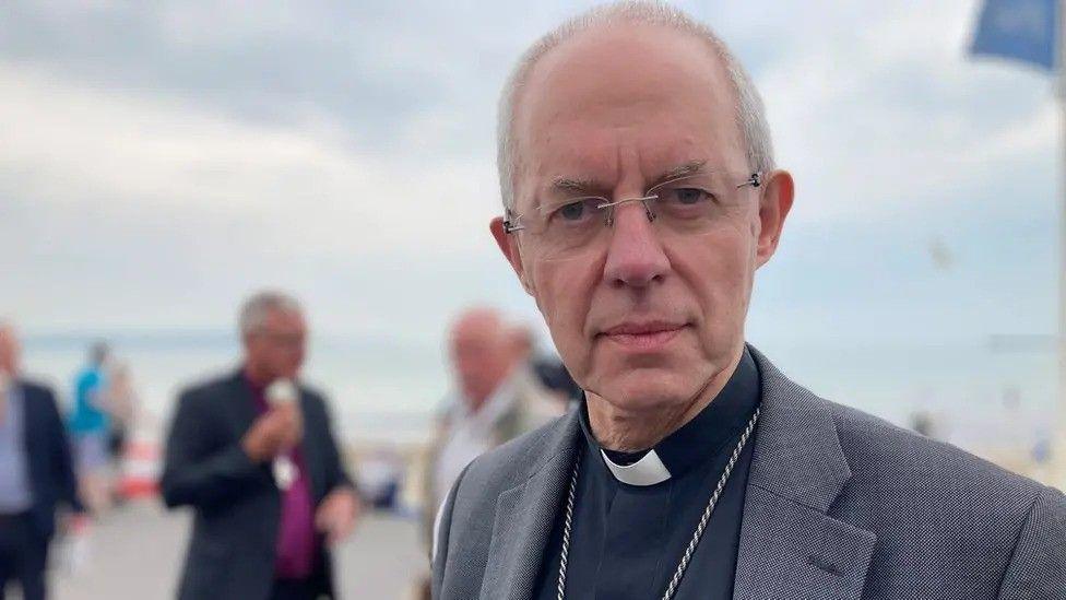 The Archbishop of Conturbury with grey hair wearing glasses, a grey suit and a black shirt with a dog collar, which is an item of Christian clerical clothing.