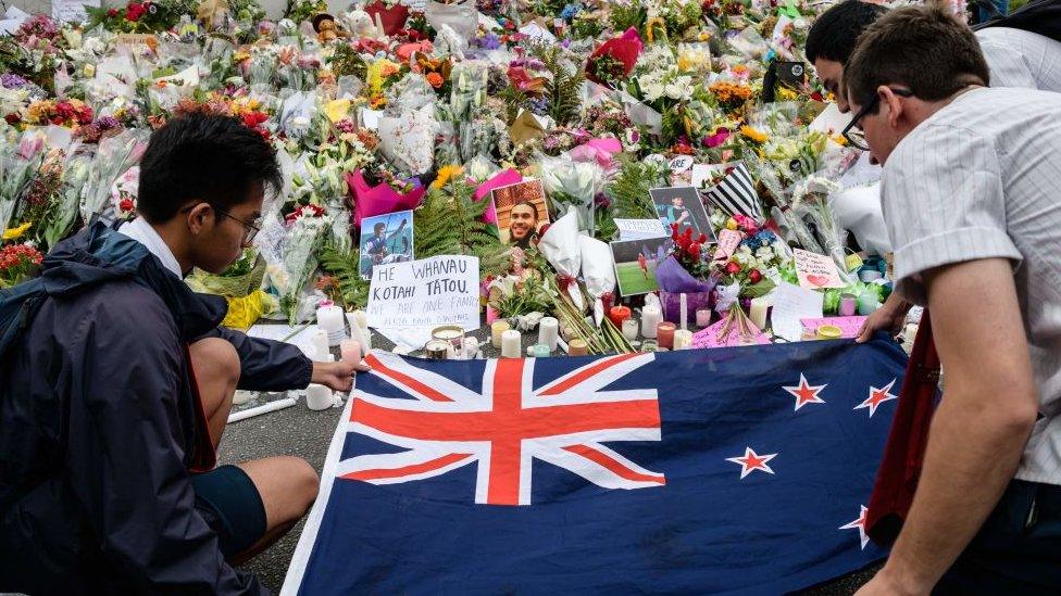 Mourners lay out a New Zealand flag next to a bed of floral tributes for Christchurch shooting victims in 2019