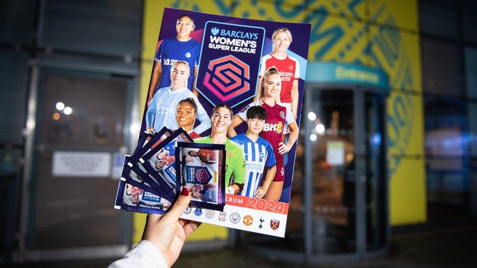 The Women's Super League sticker book being held up in front of a building. It's got seven players on the cover arranged around the Barclays Women's Super League logo. The hand holding up the album also has five packets of stickers, arranged in a fan shape, pressed against the cover with their thumb.