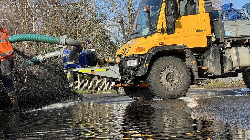 A specialist truck next to a puddle with long pipes and equipment going into the hedge at the side of a road