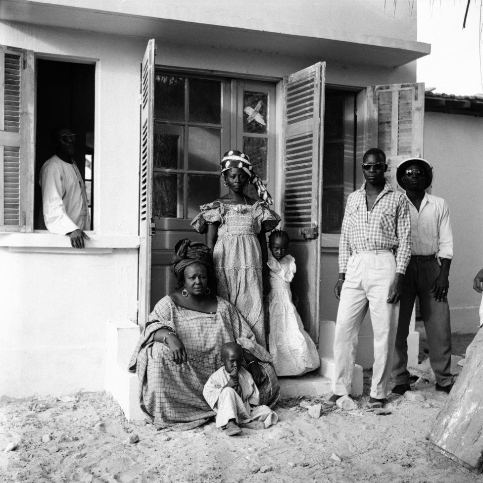 A elder woman sits at the entrance to a home, alongside infants, children and young adults.