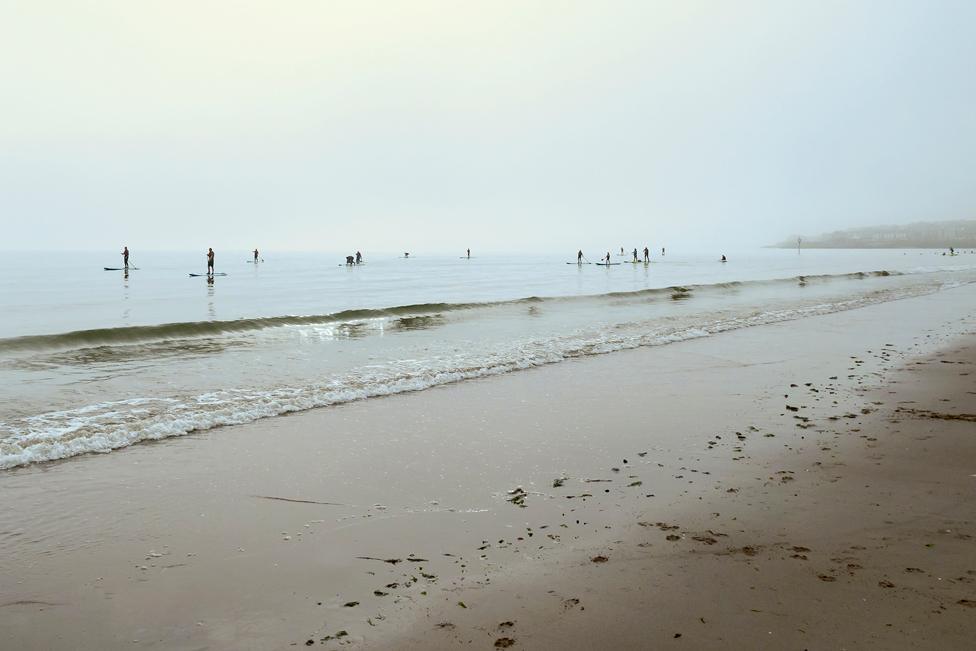 Misty Morning Paddleboarders - a sandy beach with paddleboarders floating on the sea in the distance