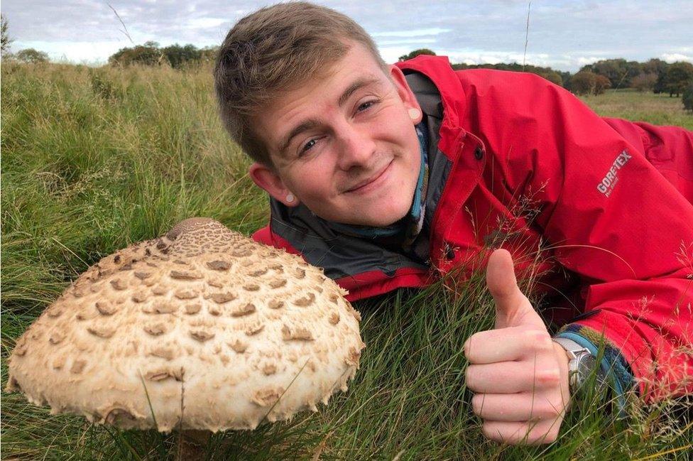 A man in a red coat gives a thumbs up while lying next to a large mushroom