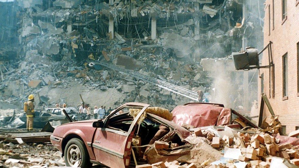 A fireman looks up at exposed floors of Murrah building, with rescuers and smashed car also pictured