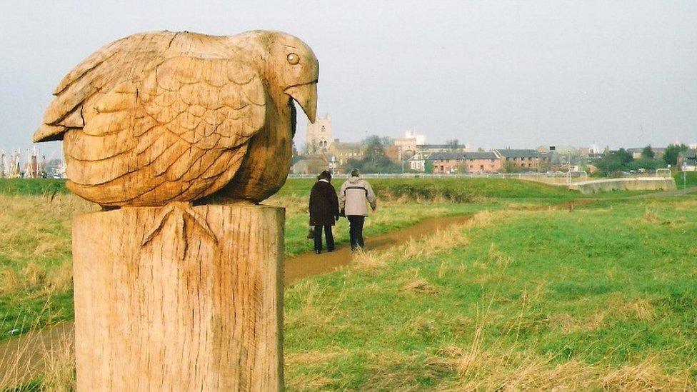 View of bird sculpture, two people walking on path across green space and King's Lynn in background