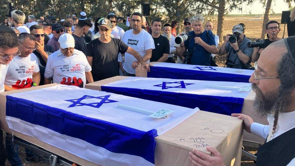 Three coffins draped in Israel flags, surrounded by mourners and journalists