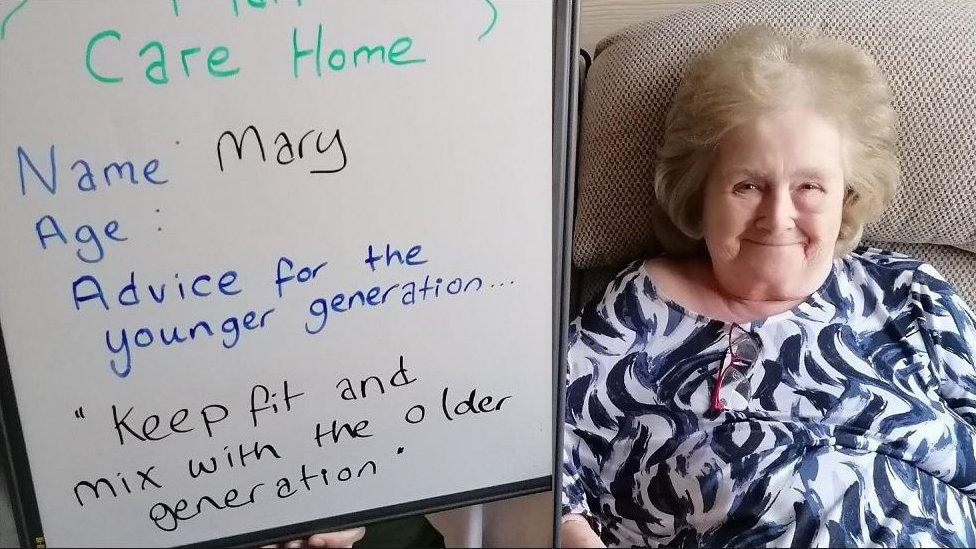 Mary is smiling at the camera in a blue and white top. A whiteboard is next to her with her message