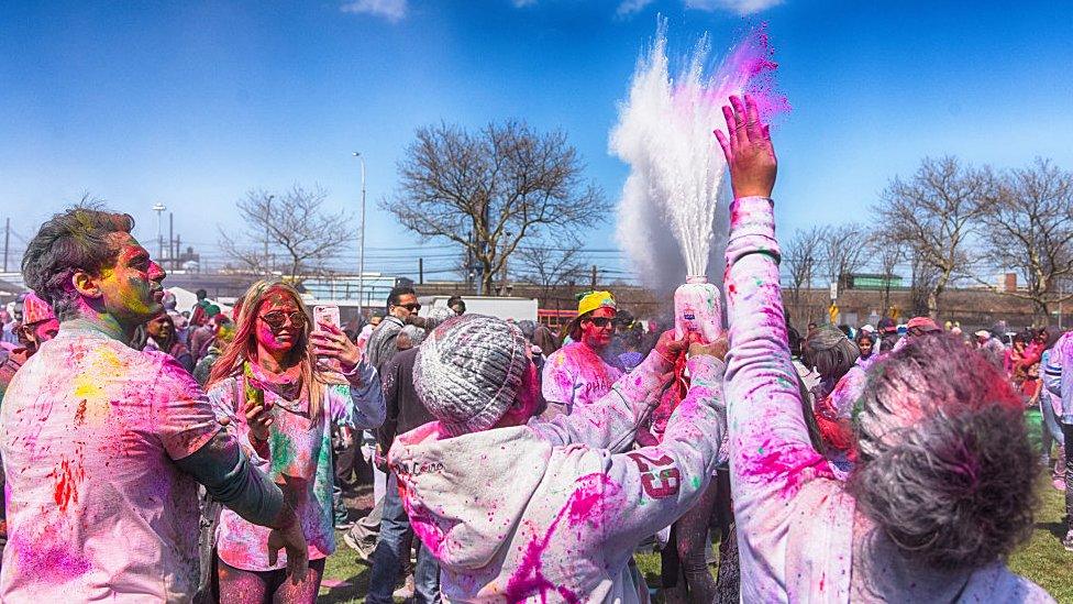 A photo from the Phagwah Festival, an Indo-Caribbean version of the Hindu Holi fest in Queens, New York. Dozens of people can be seen wearing white clothes that are completely covered in multi-coloured powders and paints. Their skin is also covered in the same multi-coloured powders and paints