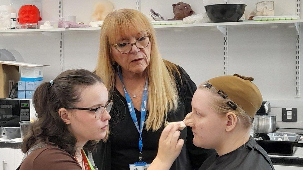 Woman with long blonde hair advises as a student with long dark hair and glasses applies make-up to a model