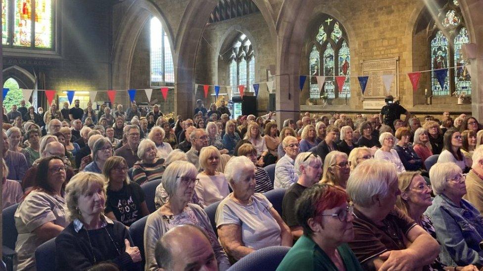 A church with stained glass windows full of people sitting down for a meeting