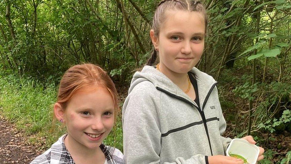 Penny (9) and Ada (10) young carers from Wiltshire standing in Jubilee Woods near Trowbridge holding tubs of items foraged from the woods