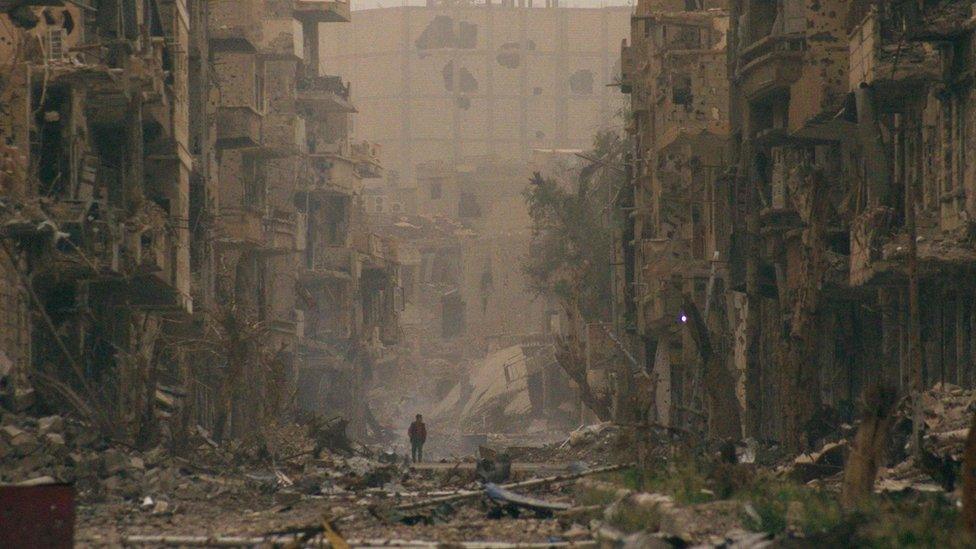 A boy stands beside damaged buildings on a street in Deir al-Zour, Syria (5 April 2013)