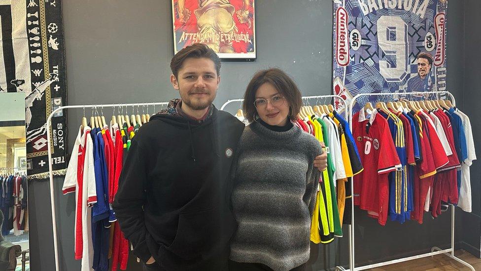 Polina Vynohradova and Serge Shcherbyna stood in their vintage shirt shop smiling for the camera. Serge is on the left and has medium length brunette hair pushed over to one side and is wearing a black hoodie. He has a slight beard and moustache. He has his left arm around Polina who is slightly shorter than him and has a brunette bob hair style and glasses and hooped earrings. She is wearing a woolly grey, black and white jumper. Behind them are three racks full of different football shirts and a grey wall.