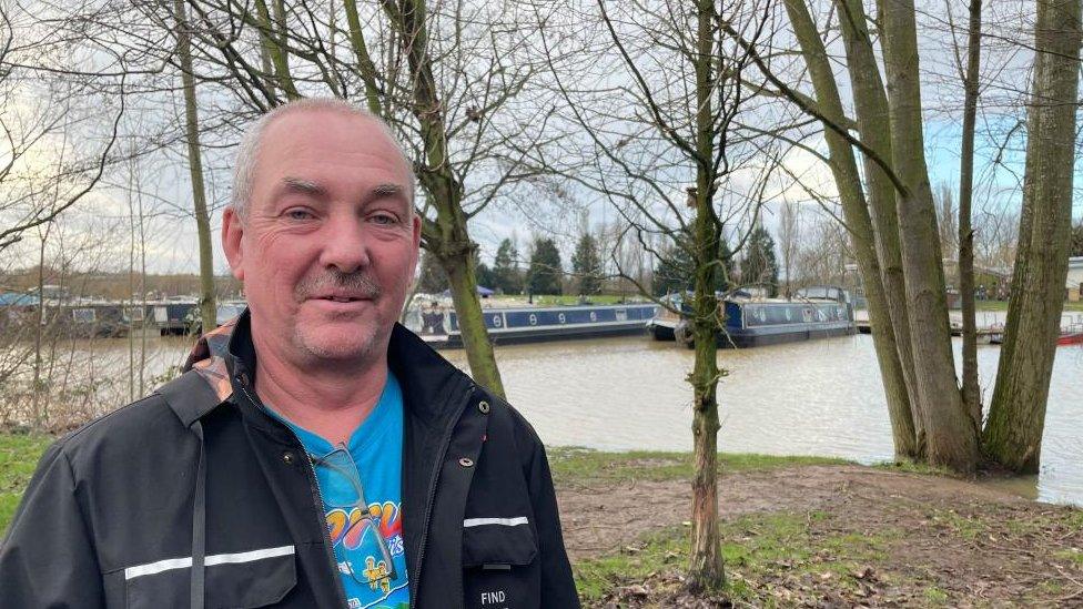 Man with short white hair wearing dark jacket stands next to a river. Narrowboats are visible.