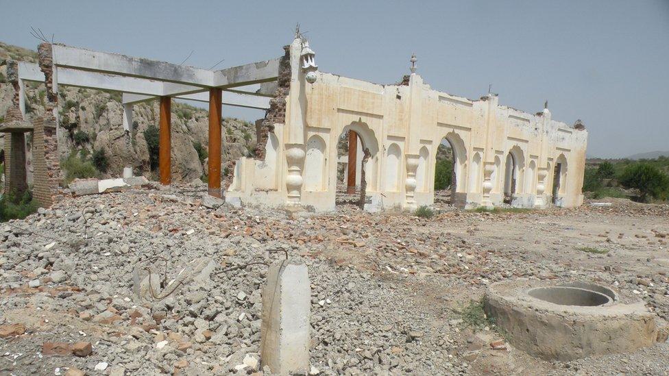 A ruined building, of which just a few beams and one wall remains, is seen on a field of rubble