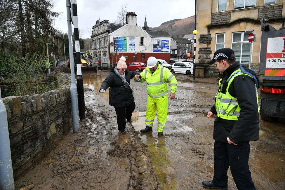 A local resident, escorted through the debris left by receding floodwater in Mountain Ash, Wales