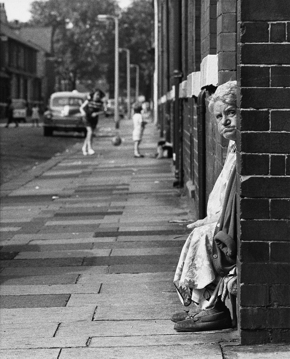 An elderly woman sits on her doorstep and looks to the camera