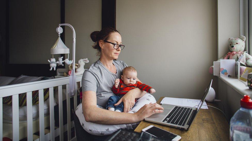 Woman working from home at a desk in her bedroom. She is using a laptop while holding her baby daughter