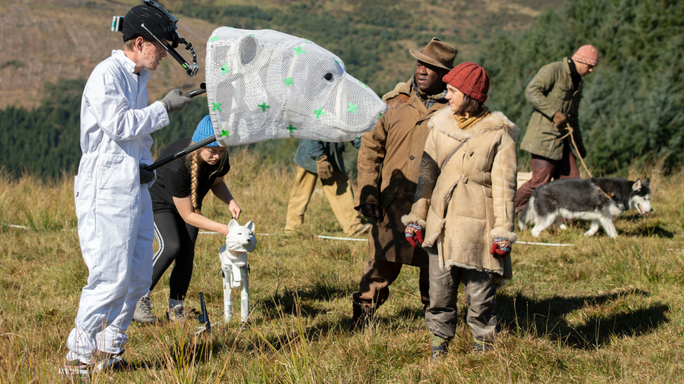 Man holds a bear-like head in front of actress Dafne Keen