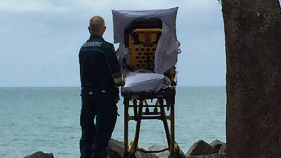 A paramedic stands behind a wheeled stretcher carrying a terminally ill woman, with both looking out at a beach