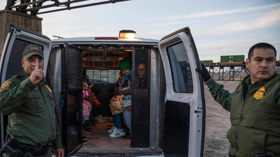 Two border staff stand by a patrol van which has a migrant family inside